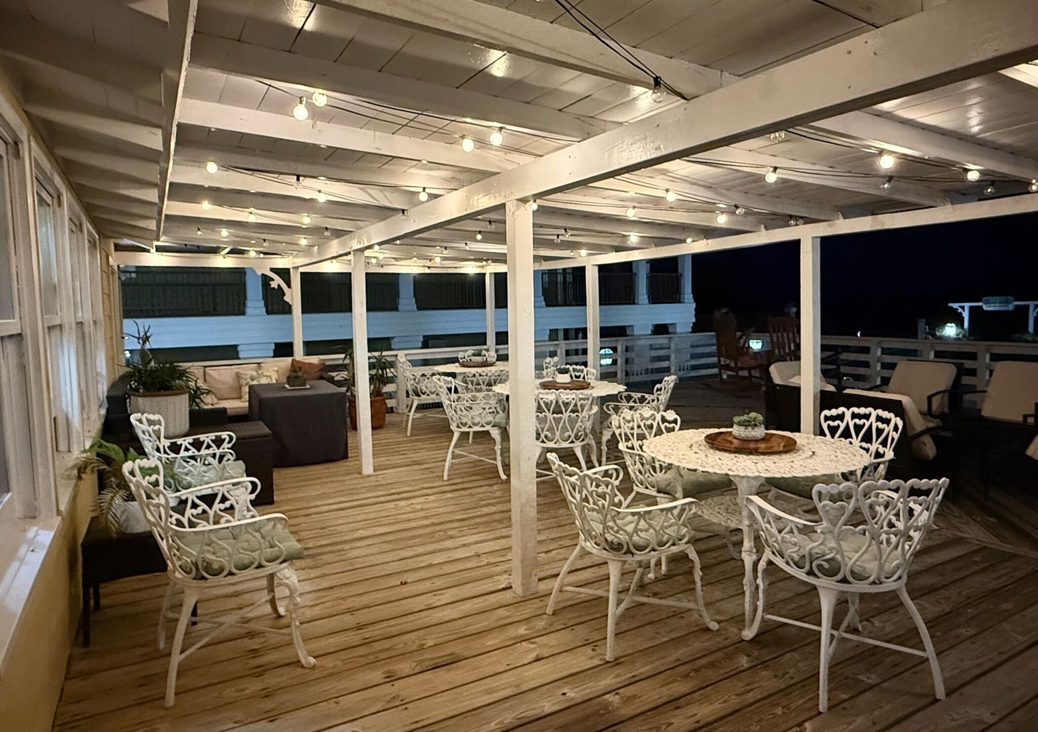 An outdoor patio with white metal chairs and tables, wooden flooring, and ceiling lights.