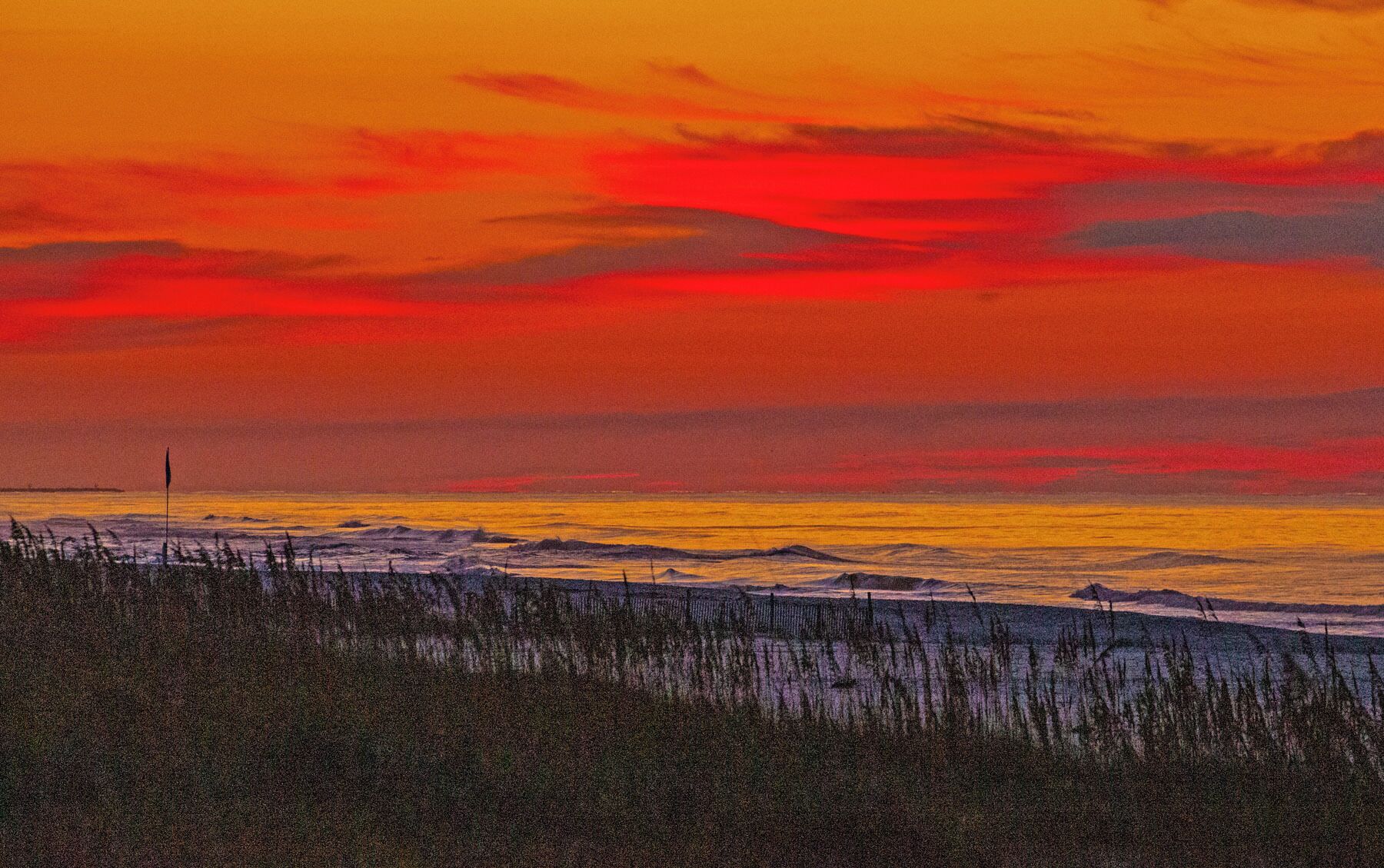 Een zonsondergang boven een strand, met golven en een vlaggenmast in de verte.