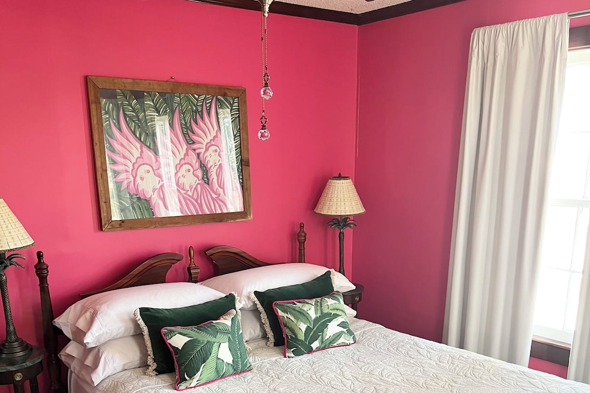 A bedroom with a bed, a ceiling fan, a painting, a lamp, and a window with curtains.
