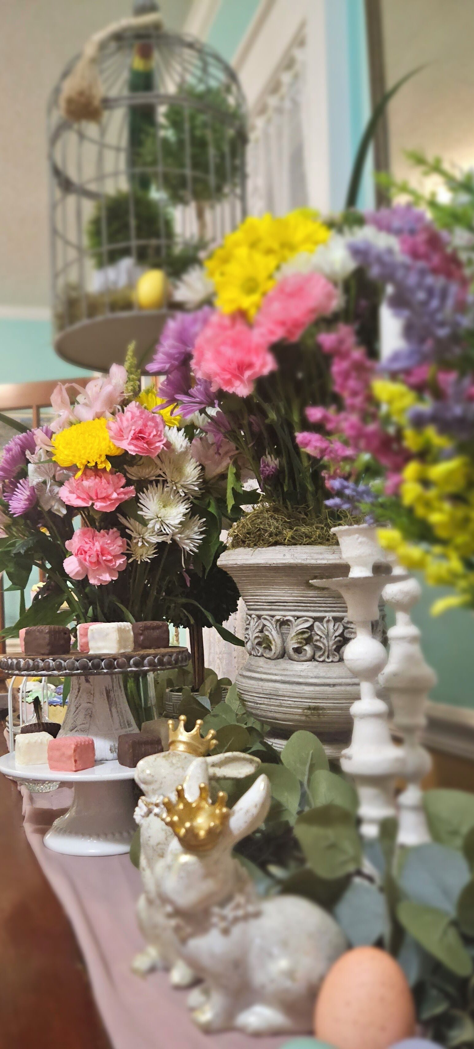 Une table sur laquelle on trouve divers objets, notamment une cage à oiseaux, des fleurs aux couleurs vives, une statuette de lapin décorative et un présentoir à étages avec des chocolats.