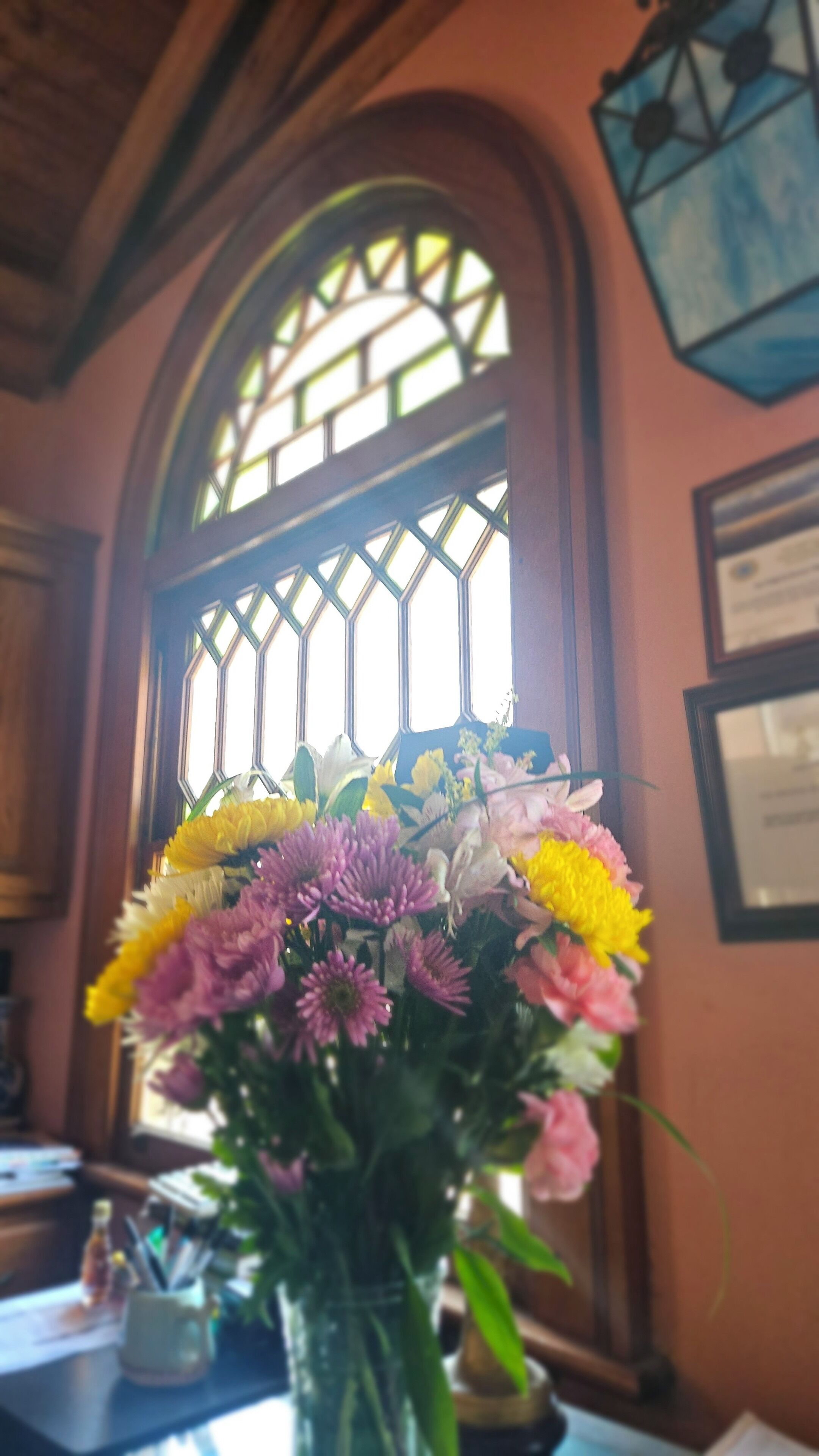 A room with a large arched window, a vase of flowers on a table, and a framed certificate on the wall.