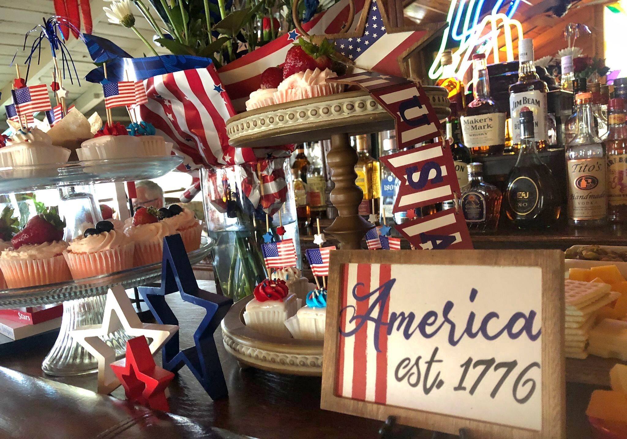 Zona de bar con decoración temática estadounidense, incluyendo cupcakes y banderas, y un letrero que dice "America est. 1776".