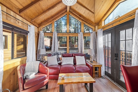 A cozy cabin interior with a wooden ceiling, a red leather sofa, a wooden coffee table, and large windows with curtains.