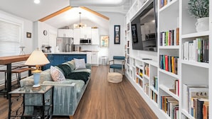 Uma sala de estar moderna com um sofá, uma mesa de centro, uma estante de livros e uma cozinha ao fundo.