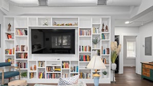 Uma sala de estar com uma grande televisão de tela plana, estantes de livros, um sofá e uma mesa de centro.