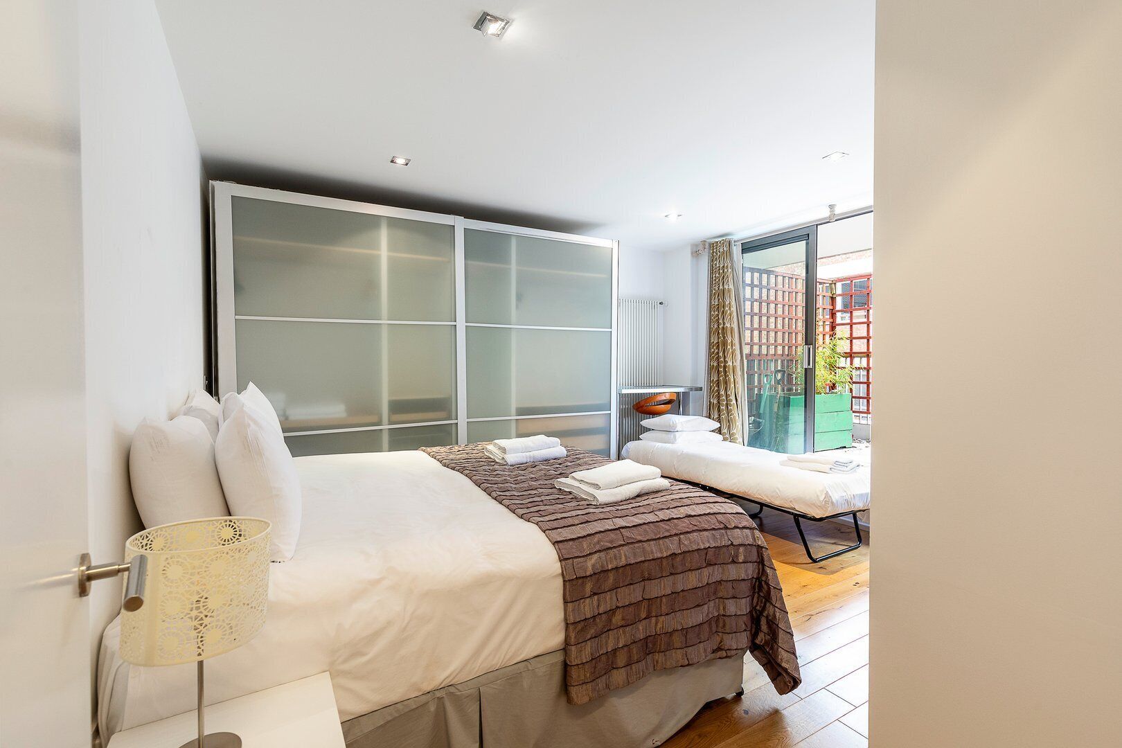 A modern bedroom with a bed, a bedside table, a large wardrobe, and a view of the outside.