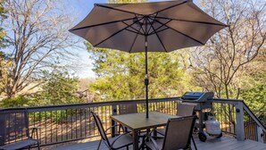 A deck with a table and chairs under a large umbrella, a grill, and a view of trees and houses in the background. - Charming Charlotte 3-bedroom house, Fenced Yard, 5 Mins to Carowinds, WiFi, Pets (Charlotte)