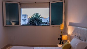 A bedroom with a bed, a nightstand with a lamp, and a window with a view of buildings and a palm tree.