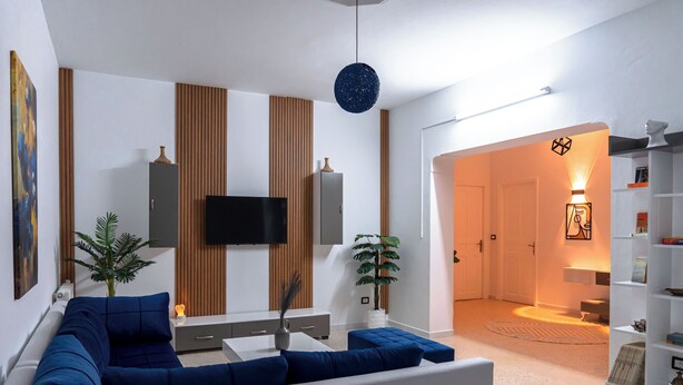 A modern living room with a sectional sofa, a flat-screen TV, wooden paneling, and a bookshelf. - CASABLU Apparte - Cozy blue terrace, Sunset view (Nabeul‎)