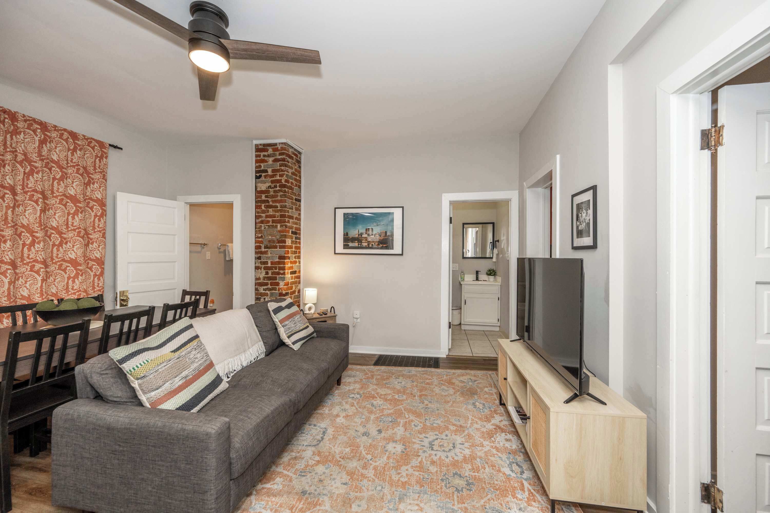 A living room with a gray sofa, a TV on a wooden stand, a patterned rug, and a brick wall.