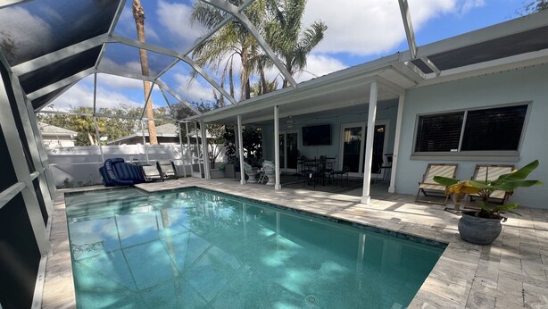A swimming pool with a tiled deck, surrounded by a covered patio area with chairs and a table.