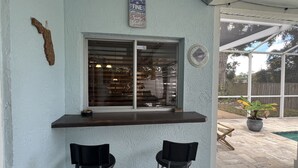 A covered outdoor seating area with a window overlooking a backyard, two black chairs, a small trash bin, and a sign on the wall.
