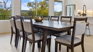 A modern dining area with a large table and chairs, large windows offering a view of the outdoors, and a kitchen area with white cabinets and appliances.
