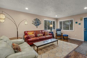 A living room with a leather sofa, a coffee table, and a chair. There are two windows with blinds, a blue door, and a decorative wall piece. - Midtown Home, Office+Fiber, Fenced Yard, Pets OK (Bend)