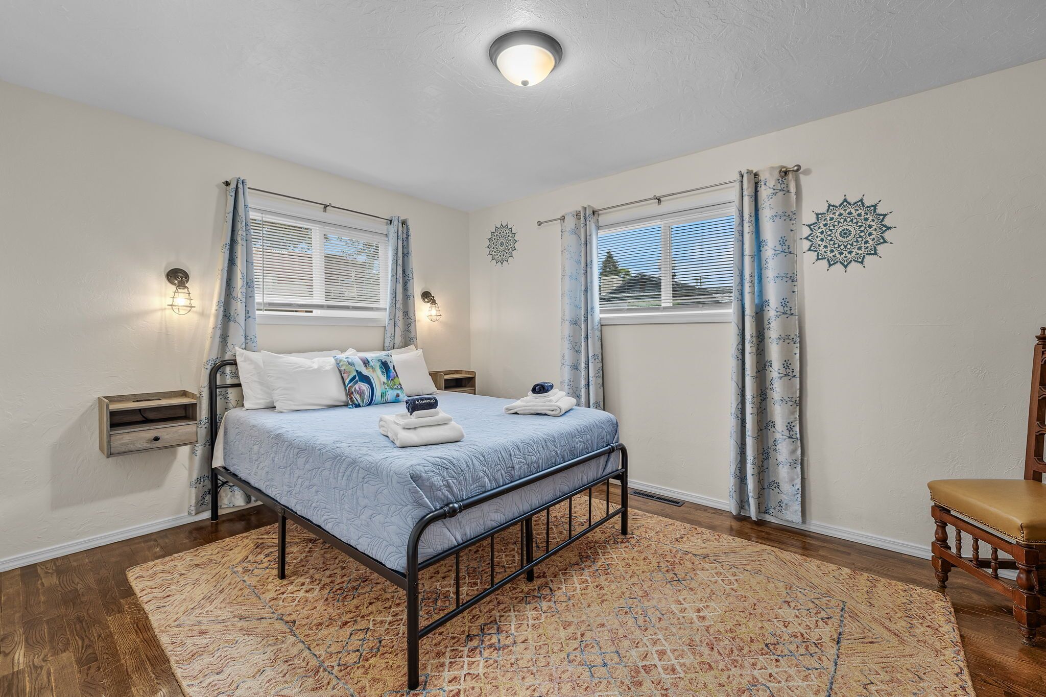 A bedroom with a bed, two bedside tables, a chair, a patterned rug, and a decorative wall piece.
