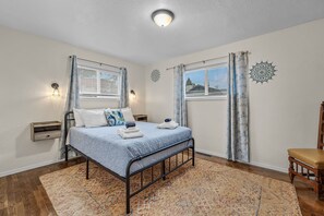A bedroom with a bed, two bedside tables, a chair, a patterned rug, and a decorative wall piece. - Midtown Home, Office+Fiber, Fenced Yard, Pets OK (Bend)