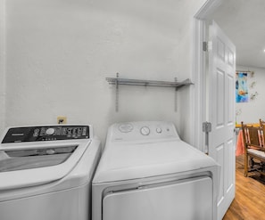 A laundry room with two front-loading washing machines, a metal shelf, and a door leading to another room.