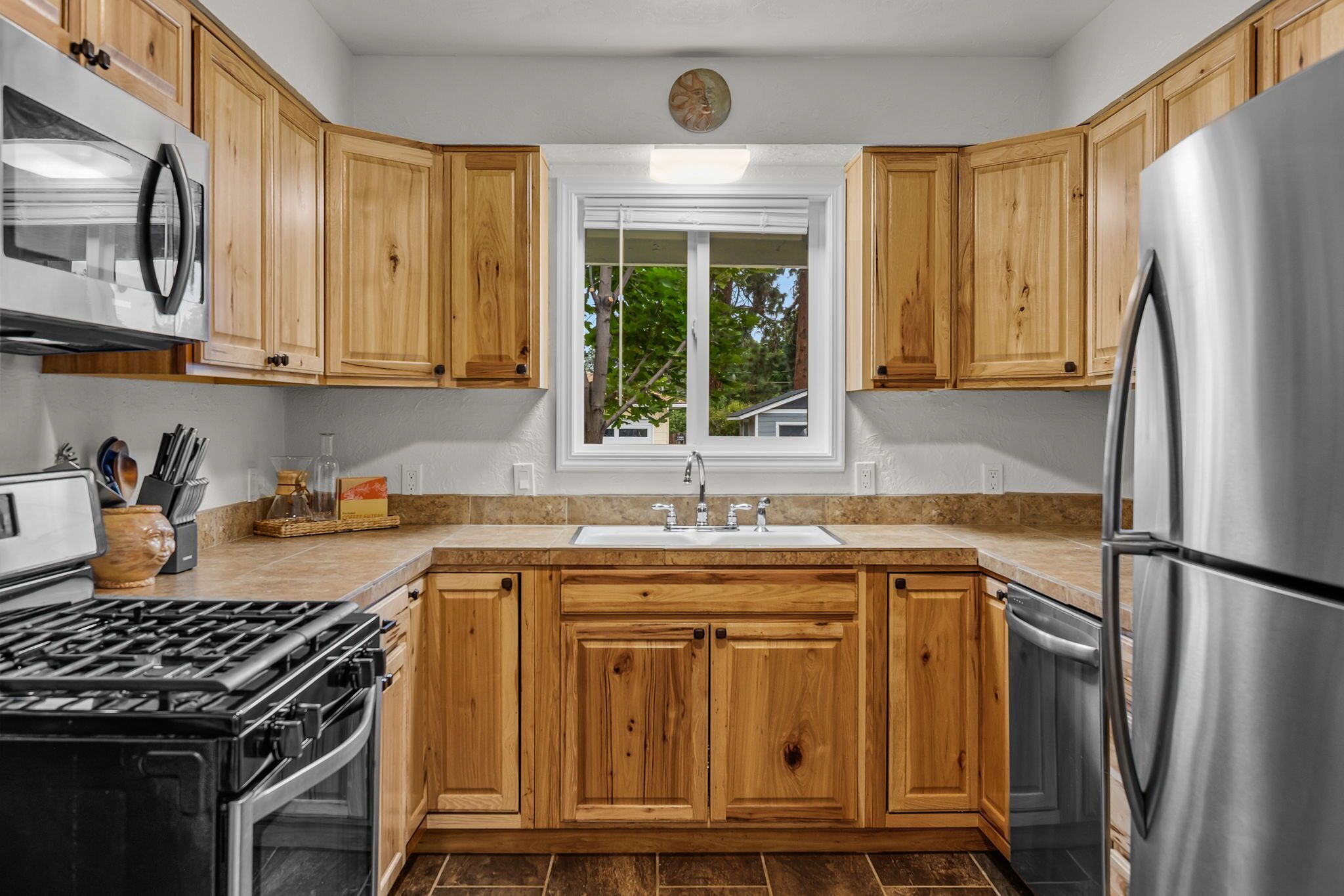 A kitchen with wooden cabinets, stainless steel appliances, and a window with a view of greenery.