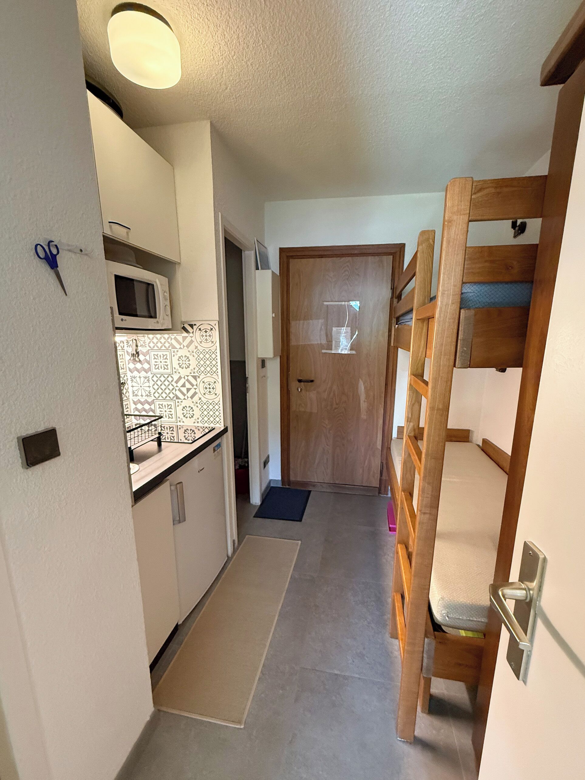 Passadís estret amb una cuineta a un costat, un llit doble de fusta a l'altre i una porta al final.