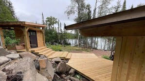 Un pontile in legno che conduce a una baita con una porta, circondata da rocce e alberi.