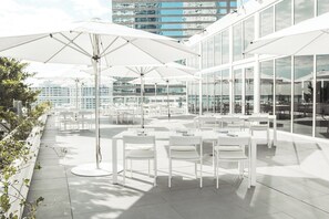 A rooftop terrace with white tables and chairs, large umbrellas, and a view of a cityscape.