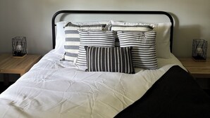 Een bed met een zwart-wit gestreept kussen, een houten nachtkastje en een aan de muur hangend beeld van surfers op het strand.