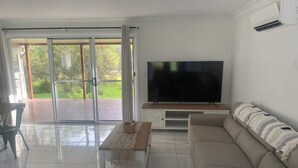 Een moderne woonkamer met een grote flatscreen televisie, een beige bank, een salontafel en een glazen schuifdeur die leidt naar een terras.
