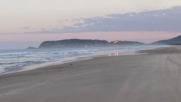 Een strand met een lange zandvlakte, golven en een verre heuvel met lichtjes.