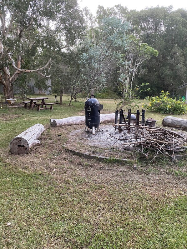 A barbecue area with a grill, picnic tables, and a fire pit surrounded by logs and greenery. - Coastal Escape (South Kempsey)