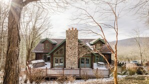 A log cabin with a stone chimney, wooden deck, and a view of the surrounding forest.