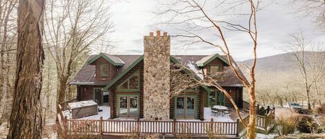 A log cabin with a stone chimney, wooden deck, and a view of the surrounding forest.