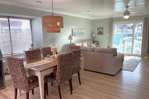 A dining area with wicker chairs and a wooden table, a living room with a sofa, a ceiling fan, and a large window with blinds.