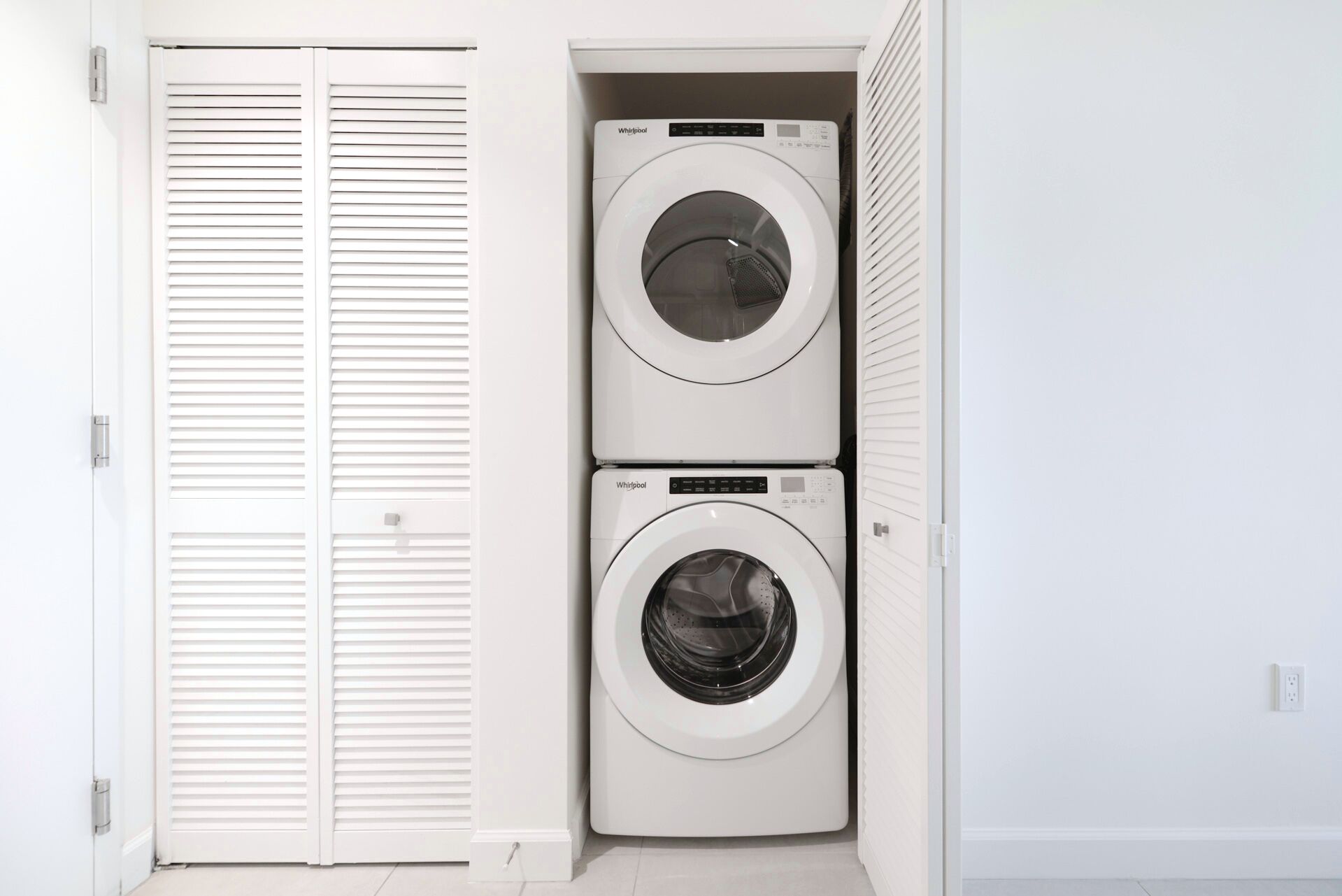 A laundry room with two stacked Whirlpool washing machines and a white louvered closet door.