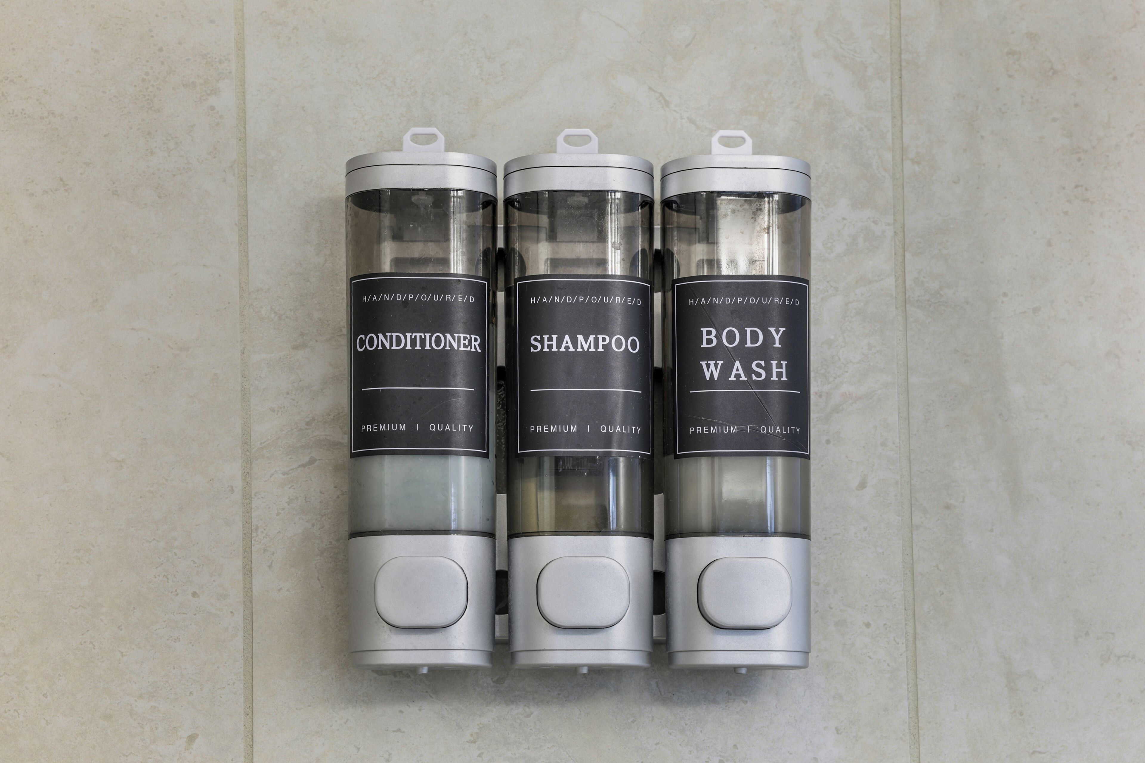 Trois récipients cylindriques, étiquetés respectivement « après-shampoing », « shampoing » et « gel douche », sont disposés sur une surface carrelée.