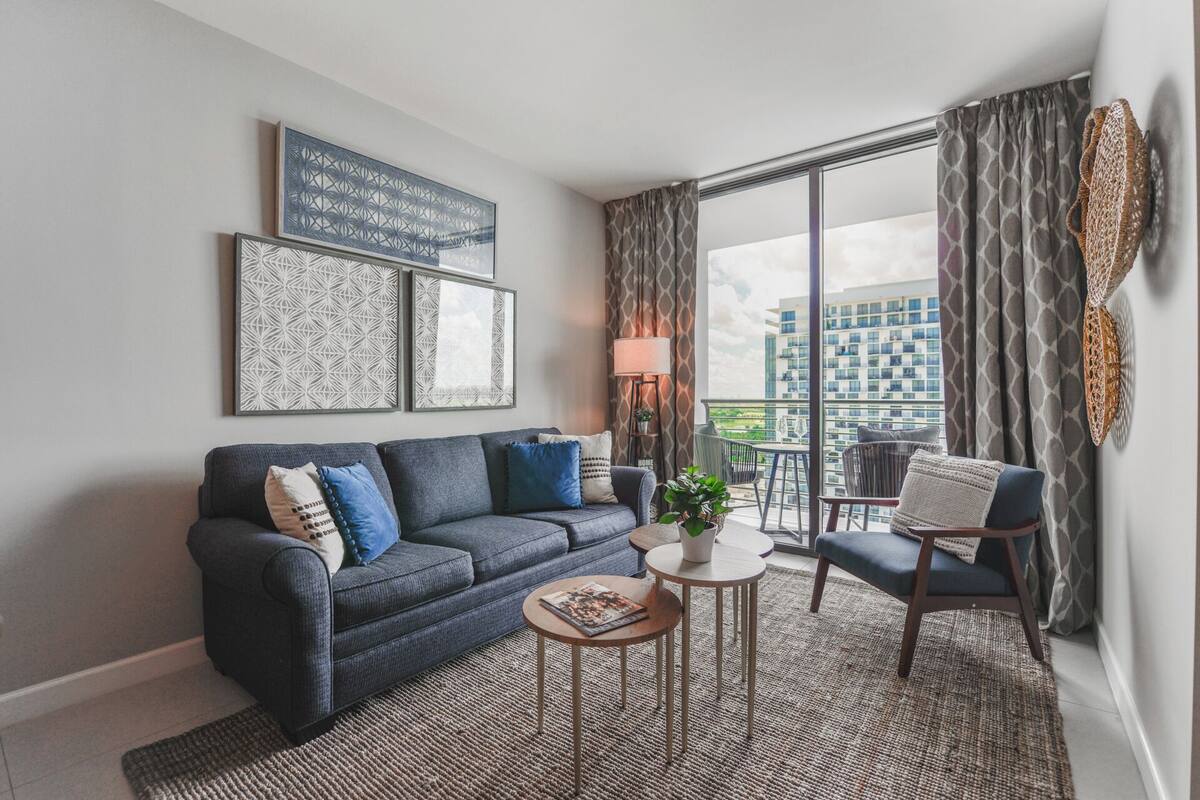 Een moderne woonkamer met een grijze bank, een klein rond tafeltje en een balkon met uitzicht op gebouwen.