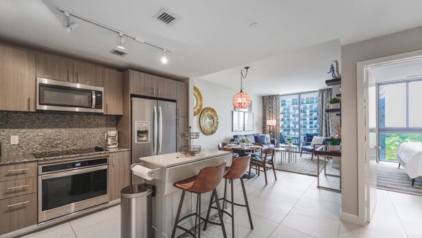 A modern kitchen with stainless steel appliances, a dining area with bar stools, and a view of a high-rise building. - 5350 Nw Downtown - Doral Building - We Host (Miami)
