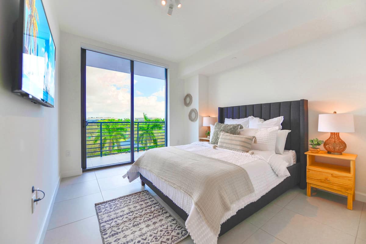 Een slaapkamer met een groot bed, een nachtkastje met lamp, een televisie aan de muur en een balkon met uitzicht op groen.