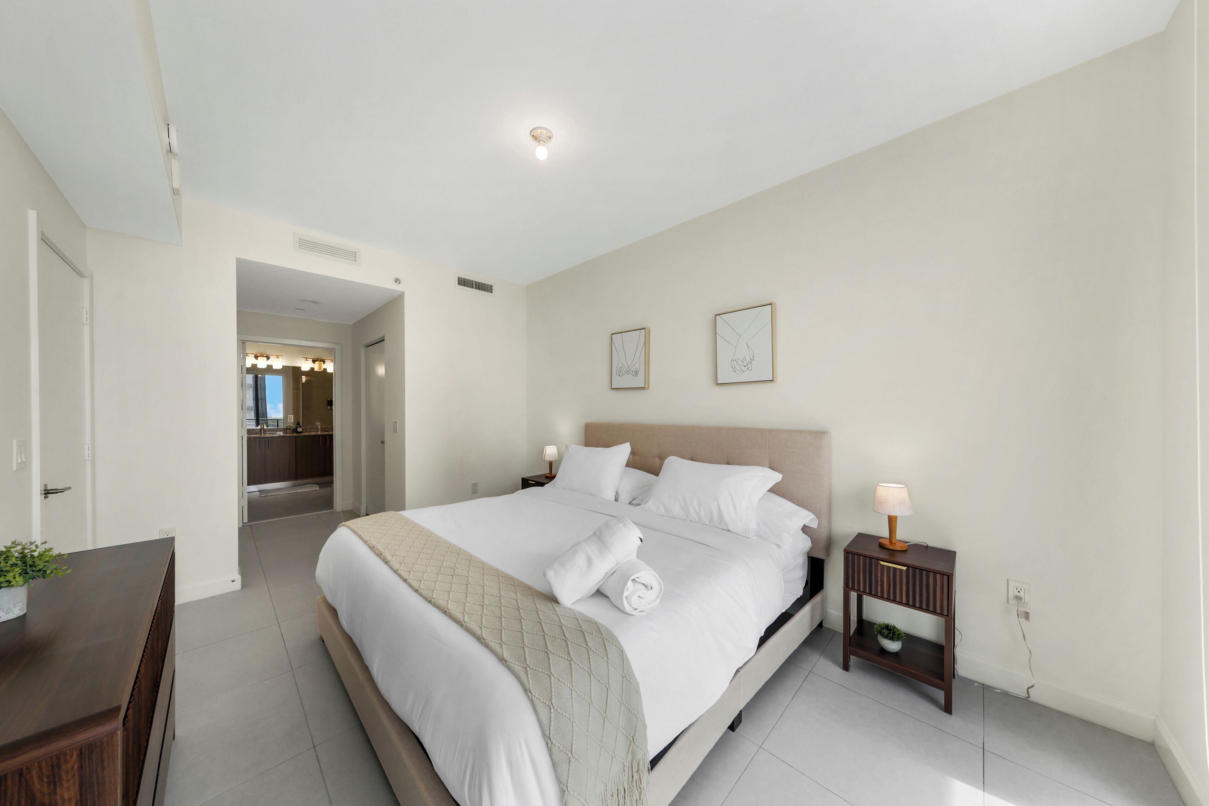 A hotel room with a large bed, a bedside table with a lamp, a small bedside plant, and a view of the bathroom.