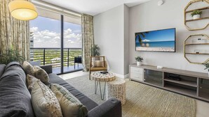 A modern living room with a large sofa, a round coffee table, and a television mounted on the wall. There is a balcony with a view of the ocean and a railing. - 5350 Nw Downtown - Doral Building - We Host (Miami)