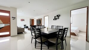 Un comedor moderno con una gran mesa rectangular y seis sillas. Se observa un sofá y una televisión al fondo.