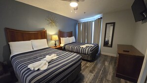 Hotellihuone, jossa on kaksi sänkyä, kattotuuletin, lamppu, peili ja puinen kaappi.