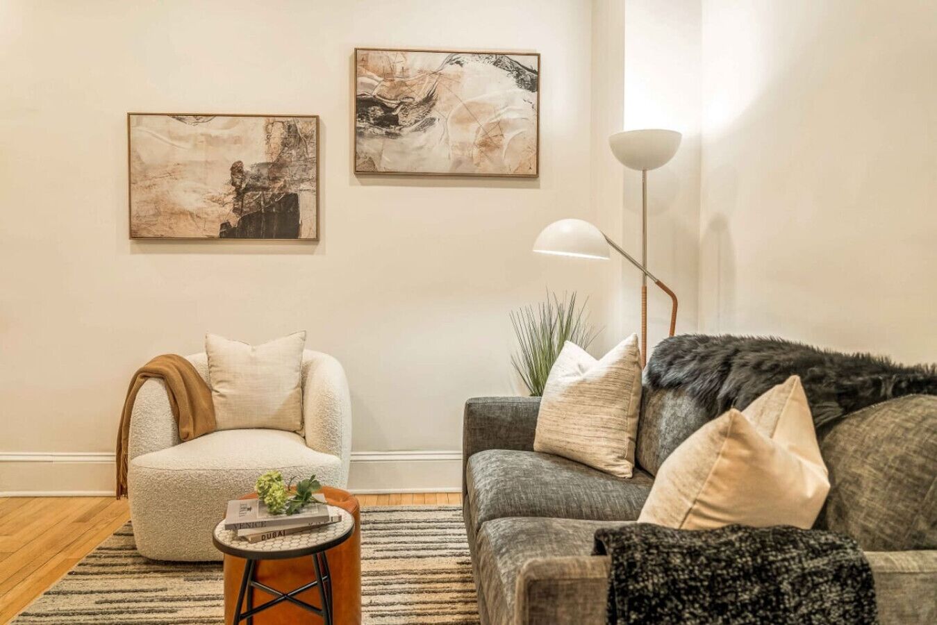 A living room with a grey sofa, a white armchair, and a coffee table with a book and a plant. There are two abstract paintings on the wall and a floor lamp.