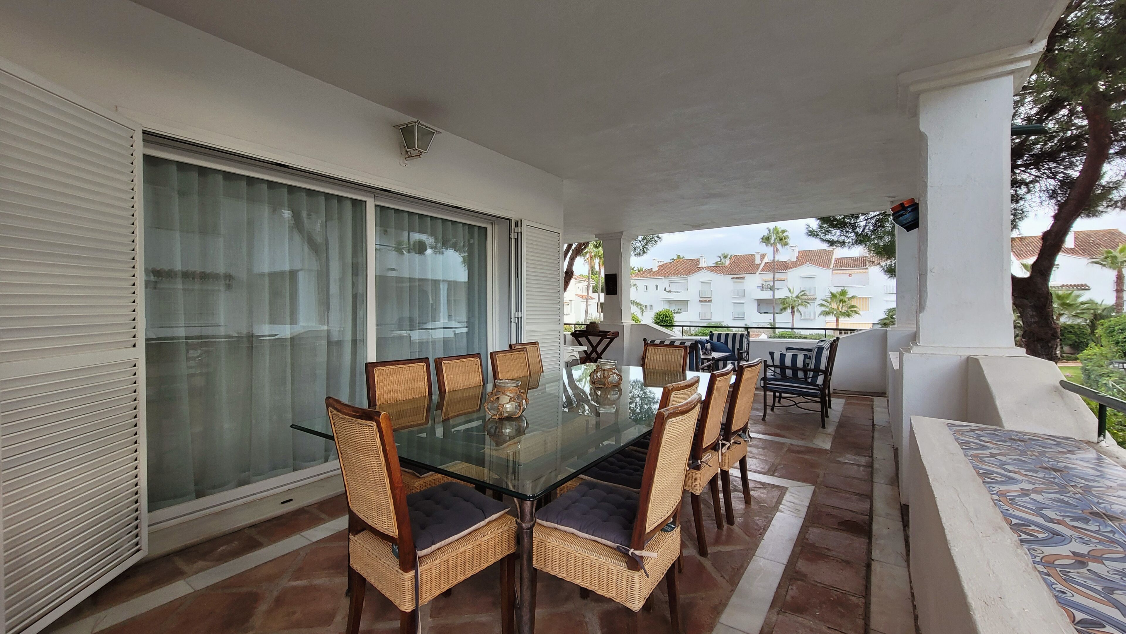 A balcony with a glass table and wicker chairs, overlooking a residential area with palm trees and white buildings.