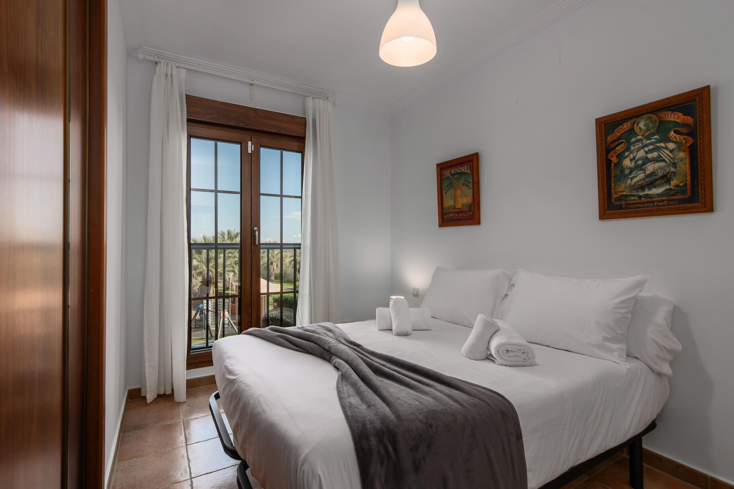 A bedroom with a bed, white curtains, a window with a view, a framed picture on the wall, and a ceiling light.