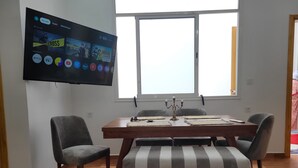 Un comedor con mesa de madera, sillas tapizadas en gris y un televisor en la pared que muestra servicios de streaming.