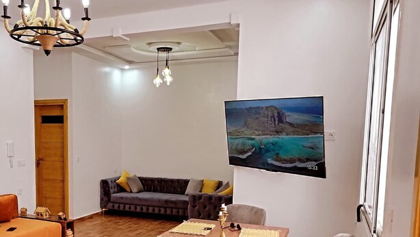 Un moderno salón con una mesa de comedor de madera, sillas acolchadas, un sofá con cojines, una araña y un cuadro de un paisaje costero.