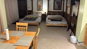 Quarto com duas camas, uma mesa de madeira e uma cadeira. Há uma janela com cortinas, um abajur e um quadro na parede.