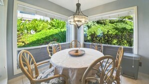 A dining area with a round table, six chairs, and a chandelier. It features large windows with a view of lush greenery.