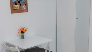 Ein kleines Zimmer mit einem weißen Tisch und zwei Stühlen. An einem Korkwand befinden sich verschiedene Gegenstände angebracht, darunter ein Schild mit der Aufschrift 'Welcome' und ein Rauchverbotsschild. Auf dem Tisch steht eine Vase mit Blumen.