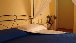 Un lit avec une couvre-lit bleue et des oreillers blancs, une table de chevet ornée d’un vase de fleurs et un rideau blanc.